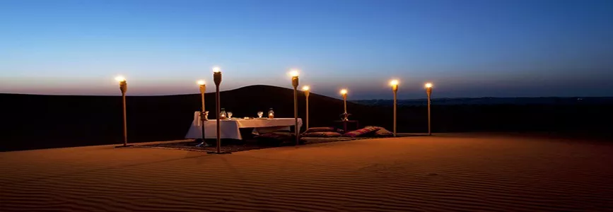 Lamp-lit dining table set on the dunes at a desert camp at dusk