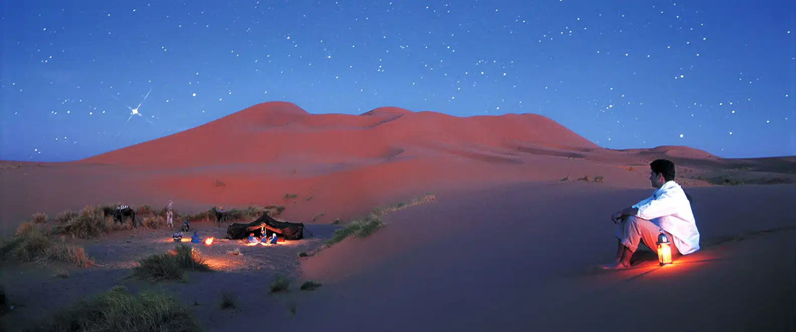 Traveller sitting by a lantern at a desert camp under a starry sky, Morocco