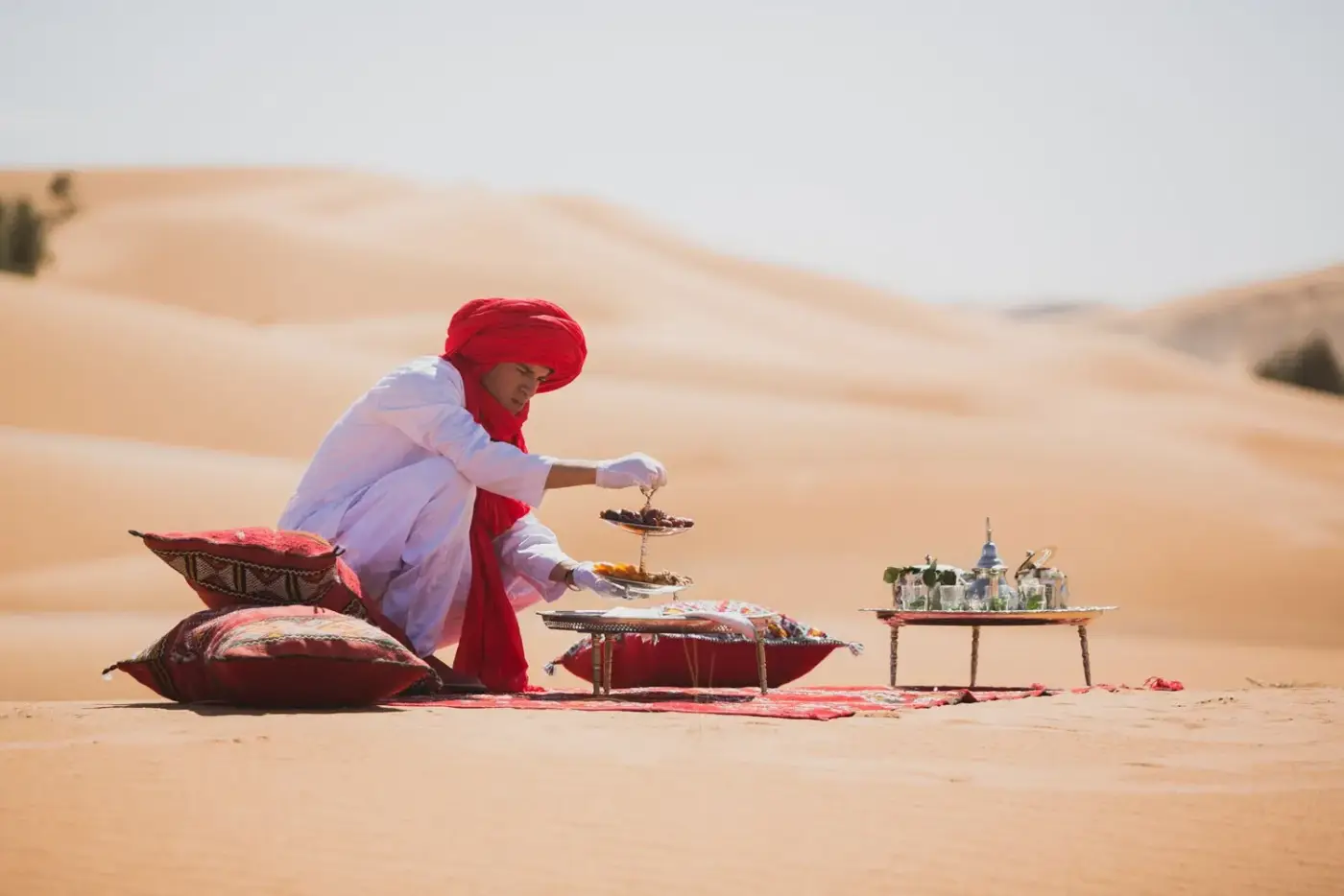 Moroccan host in a red turban serving mint tea and dates on a carpet in the Sahara dunes